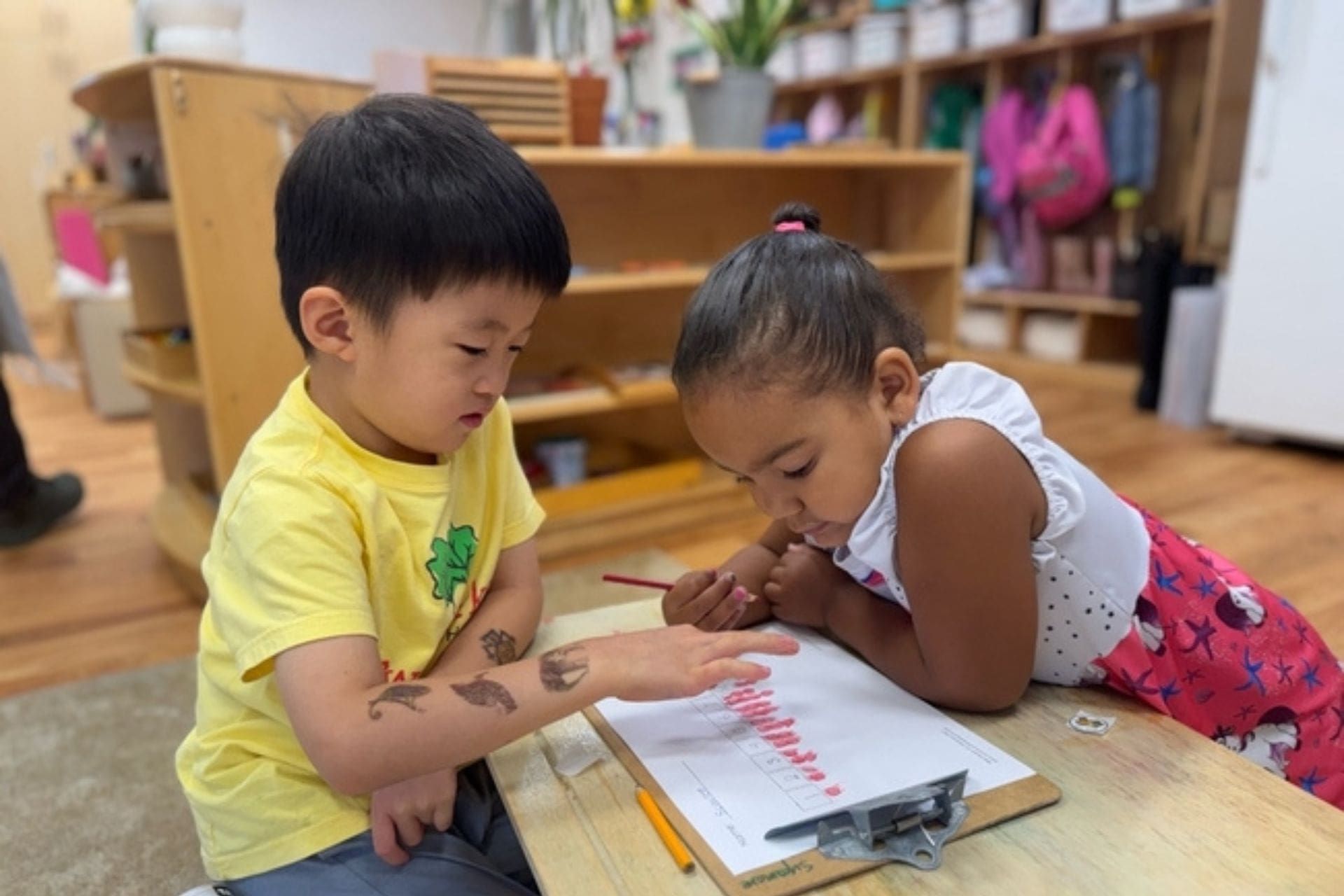 Two children, one in yellow shirt, draw on paper together at a table in a Montessori classroom.