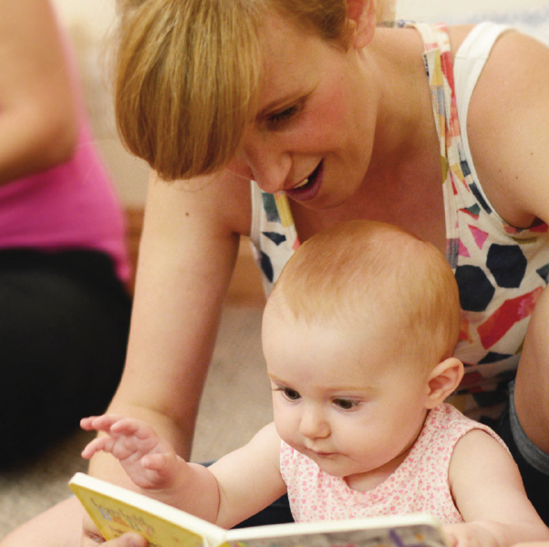 Woman reading a book with a baby, both are smiling. Indoors, soft lighting.