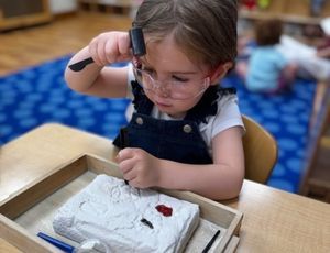 Child in safety glasses uses a tool on a plaster-like block in a tray, indoor setting.