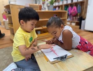 Two children at a desk, one with drawn marks on their arm, the other drawing on paper.