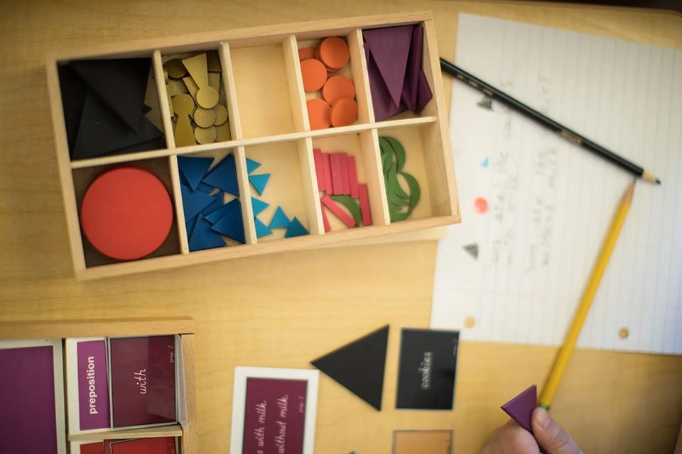 Wooden box with colored geometric shapes. Person holds a triangle next to a pencil on paper with a diagram.