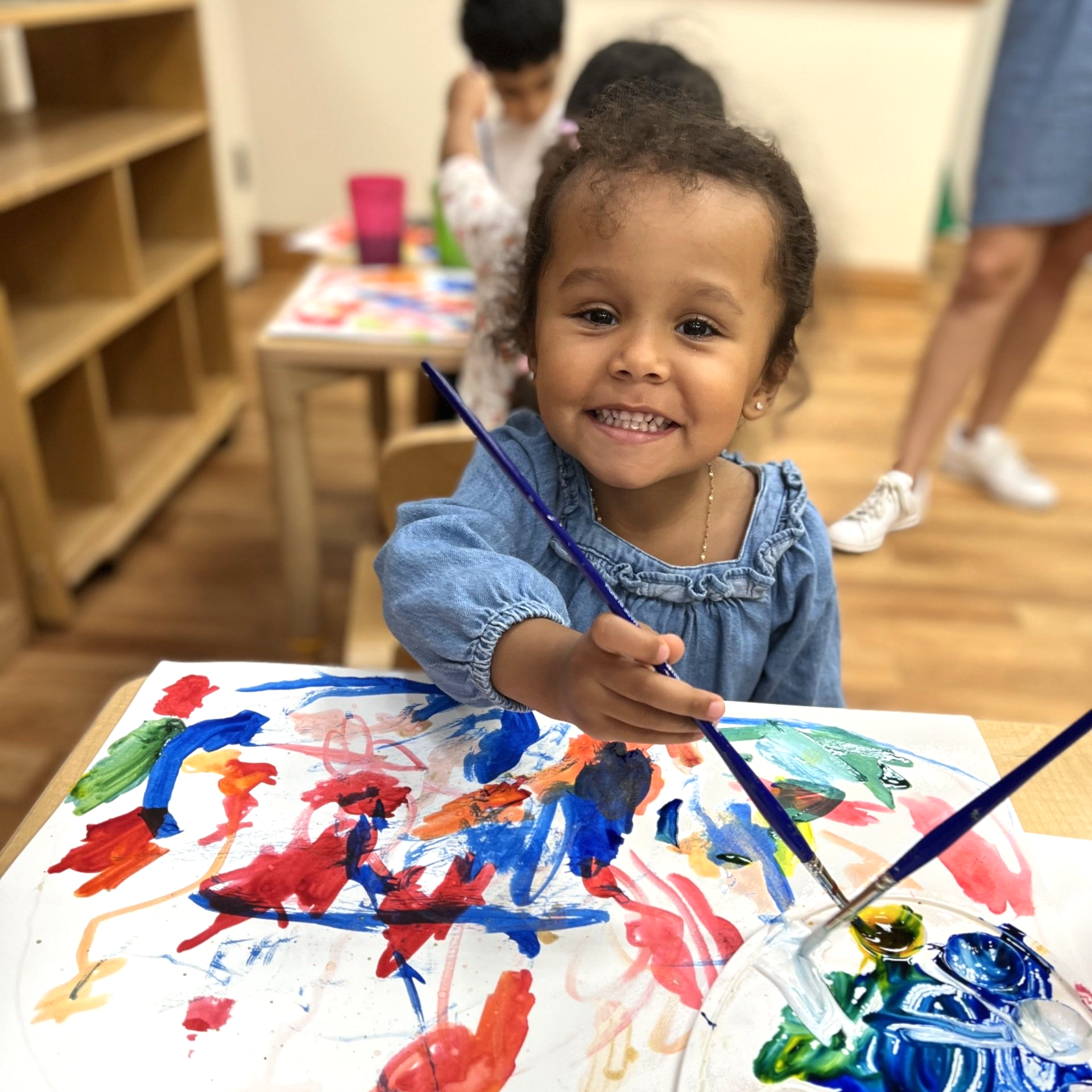 Young child painting at a table, smiling. Colorful artwork and paint palette visible. Bright classroom setting.