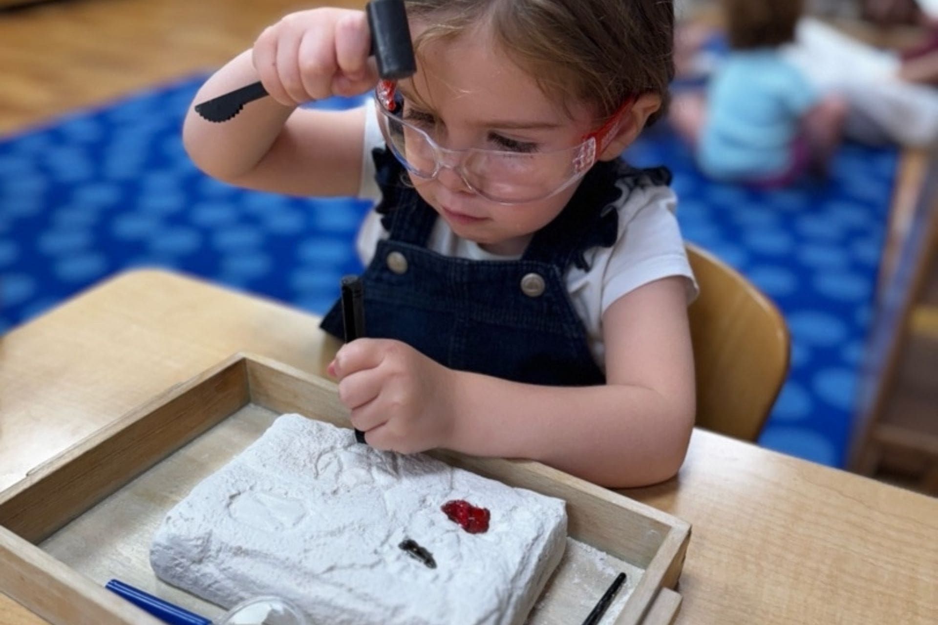 Child with safety glasses, using tools to examine an item in a tray.