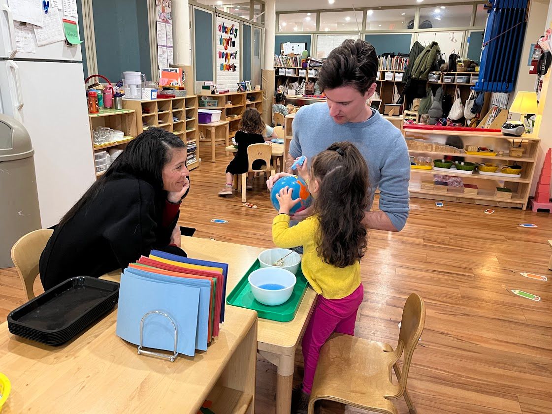 Two adults and a child playing at a table in a brightly lit classroom.