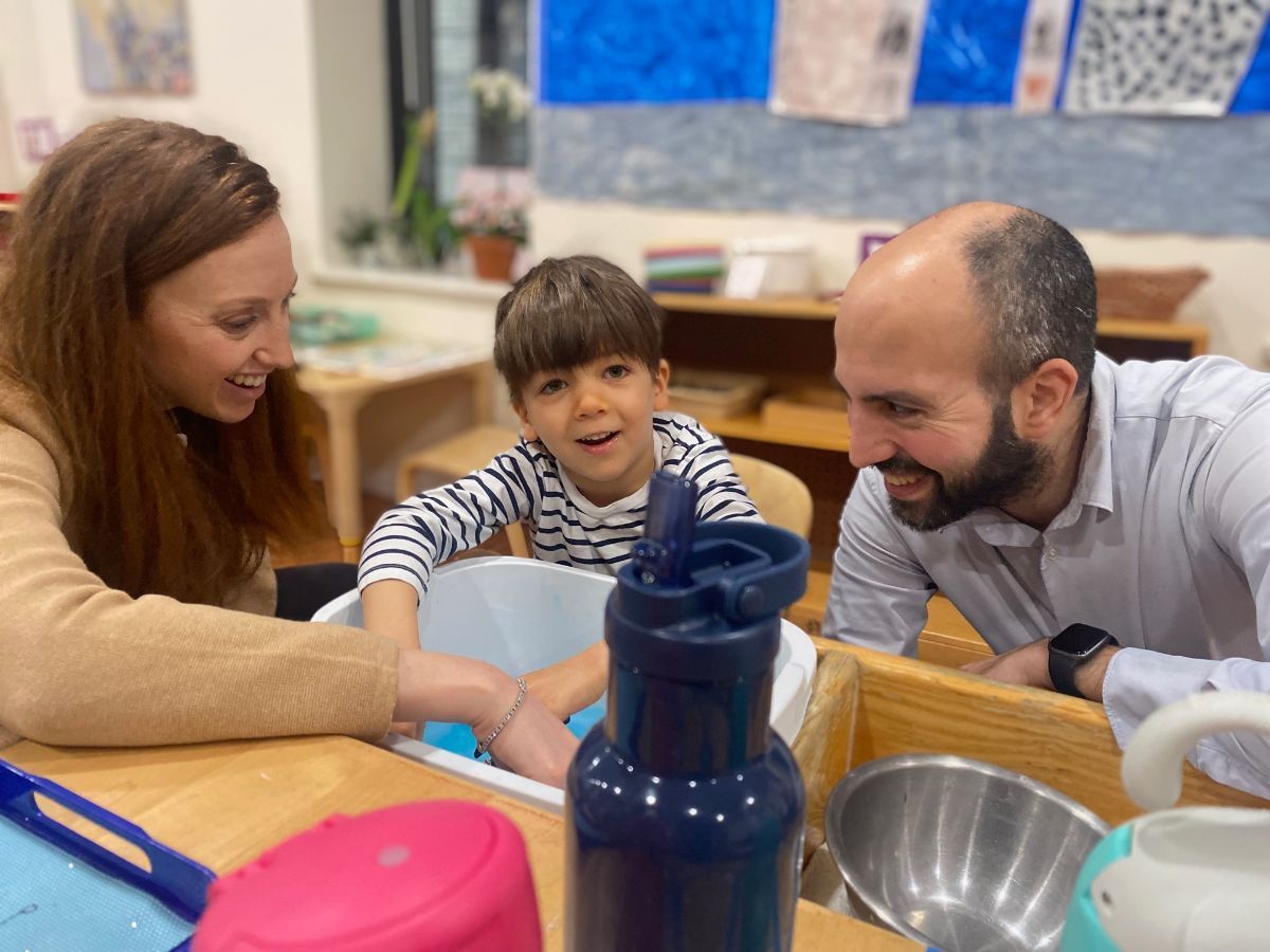 A woman and man watch a boy playing with water in a classroom setting. They are smiling.