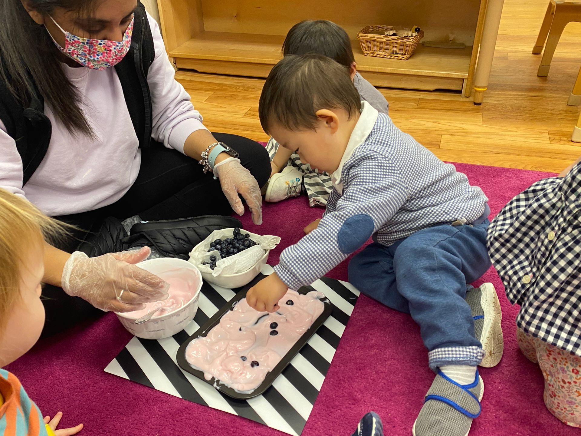 Children and an adult in a classroom, making a decorated treat. The child in front is putting blueberries on.