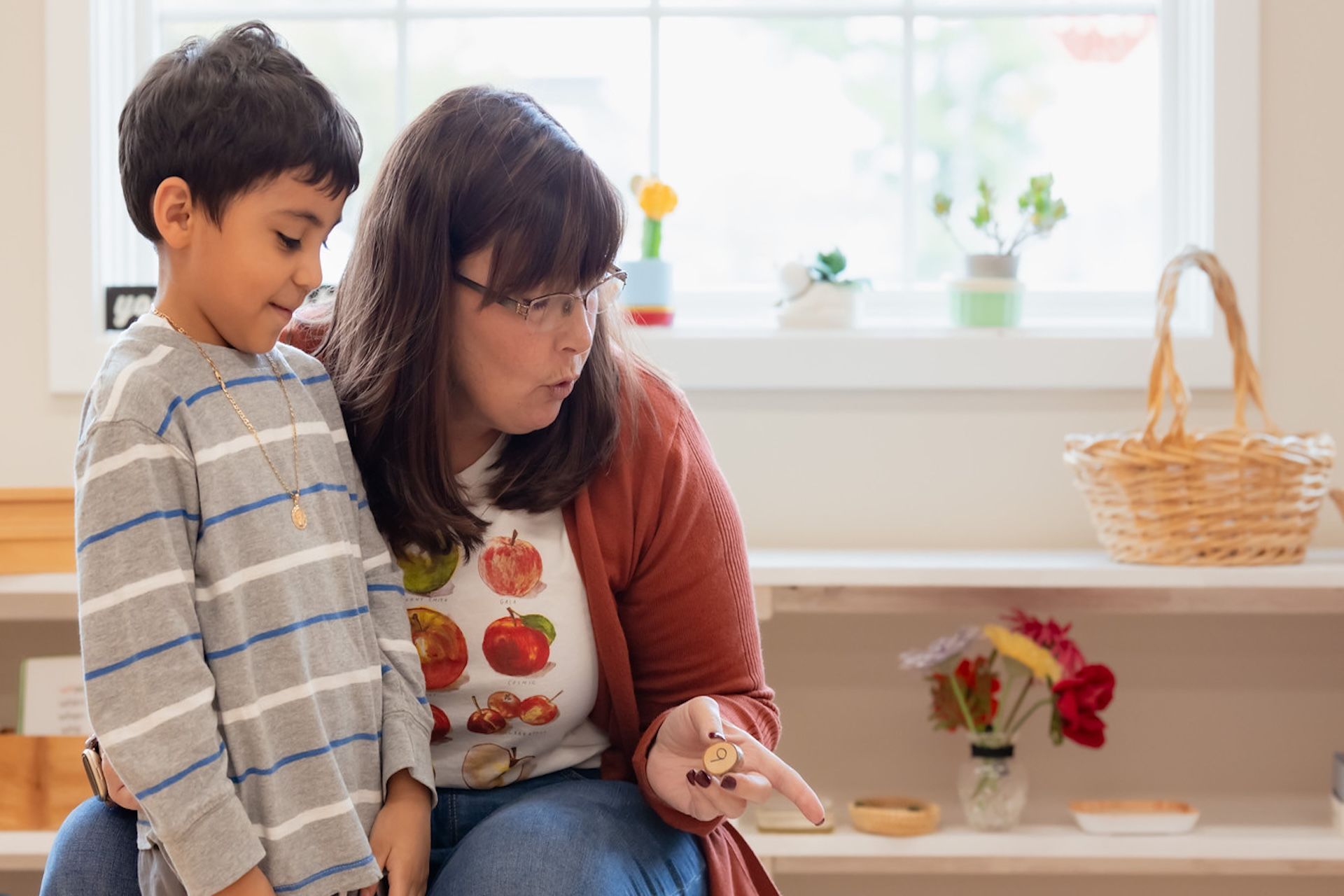 A teacher holds a small item to show a student in a bright, organized classroom setting.