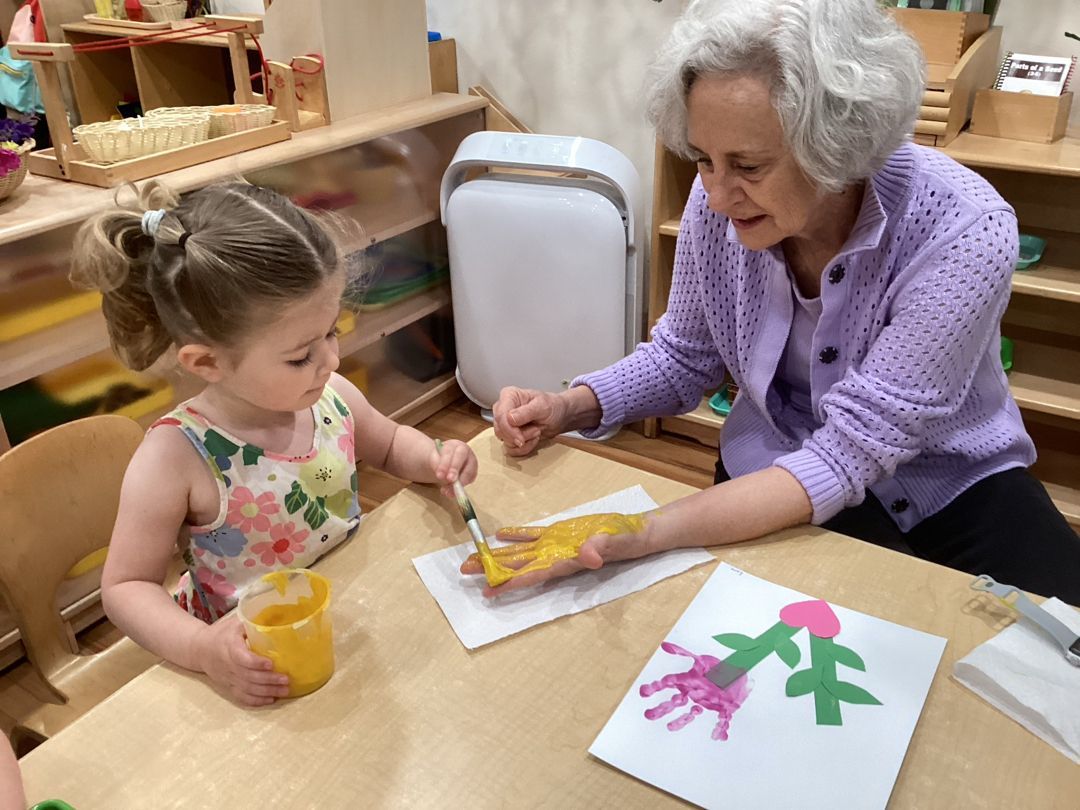 Child and adult doing handprint painting at a table indoors; child holds paint, adult has yellow-painted hand.
