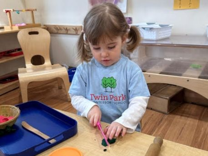 Young Montessori child cutting playdough with a pink knife at a table in a classroom.