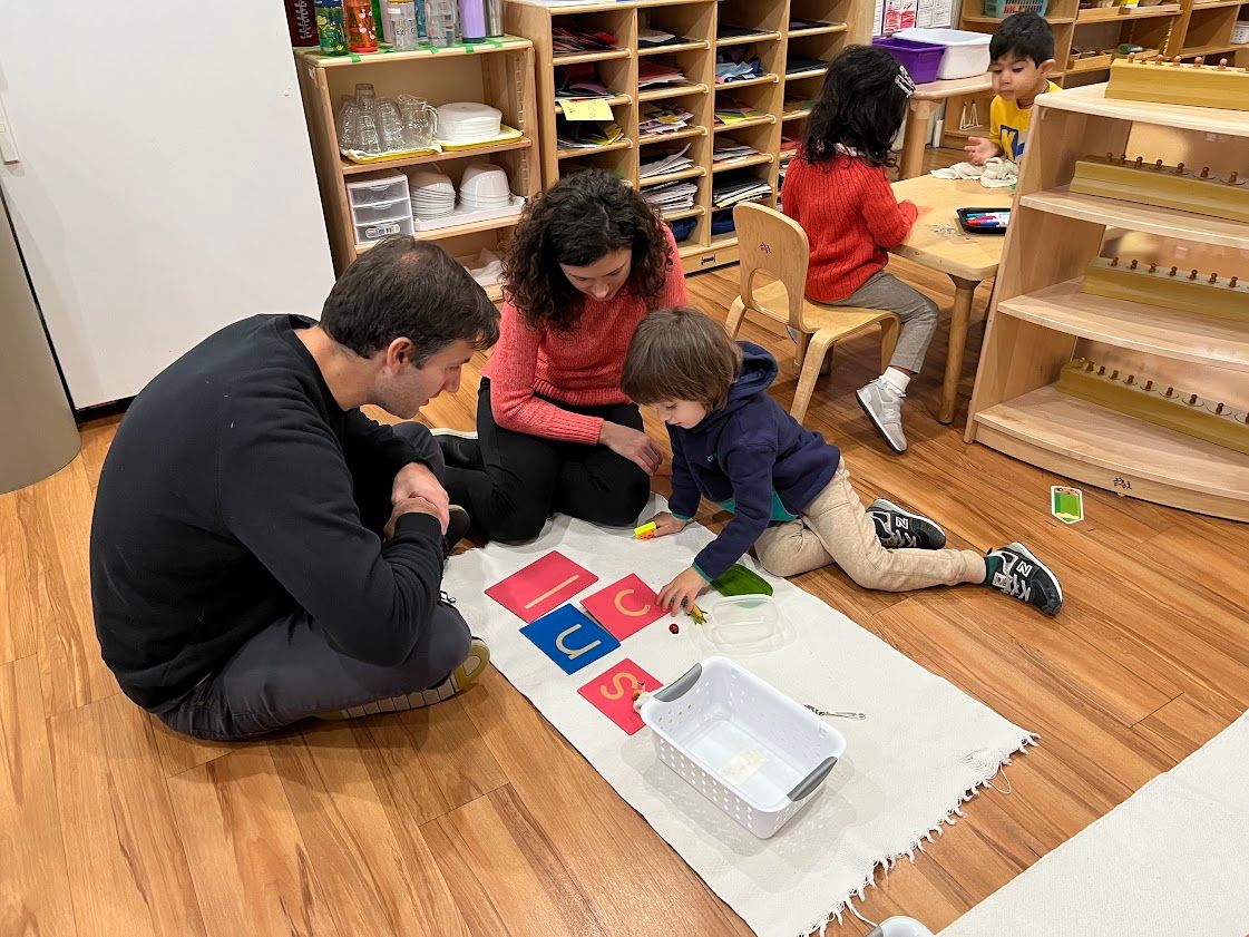 Two adults and a child playing a word game on the floor in a classroom.
