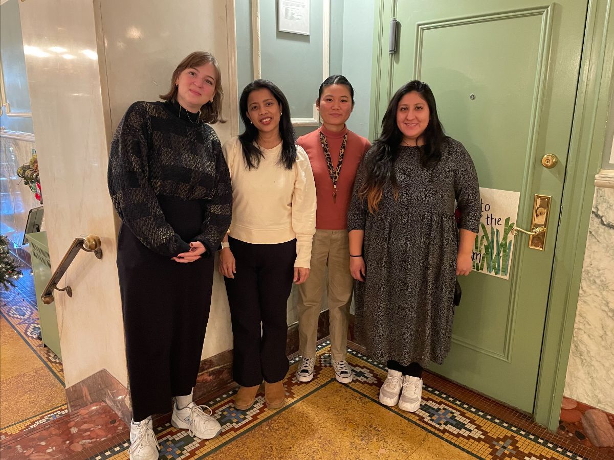 Four women stand in front of a green door. They are smiling, near a decorative floor in a hallway.