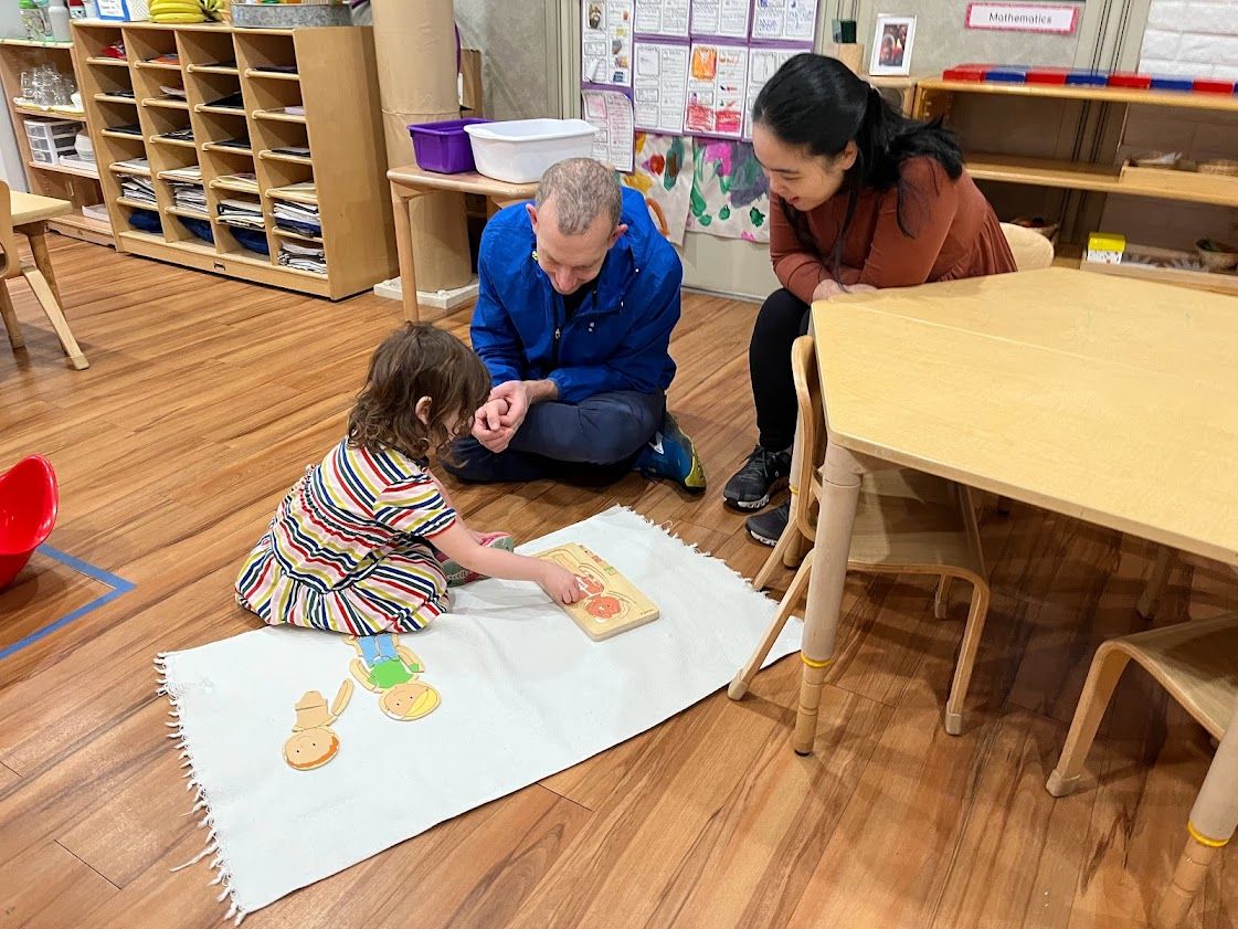 Child doing puzzle on mat, with teacher and man watching in classroom.