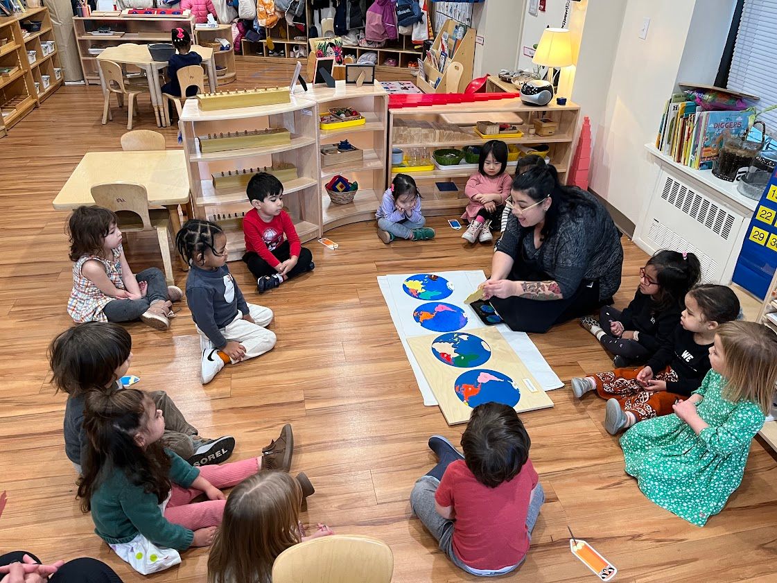 Children in a circle watch a teacher demonstrate colorful shapes on the wooden floor of a classroom.