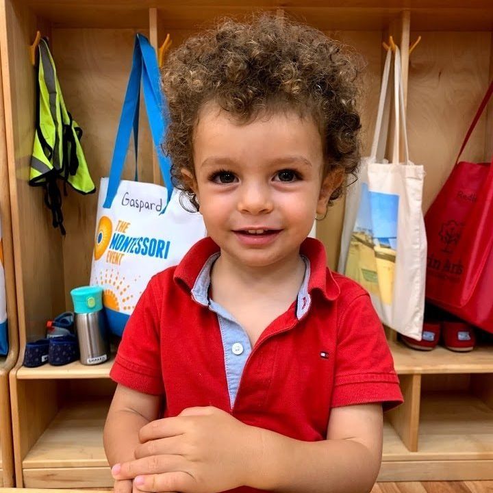 Young child with curly hair in red shirt smiles in front of wooden cubbies.
