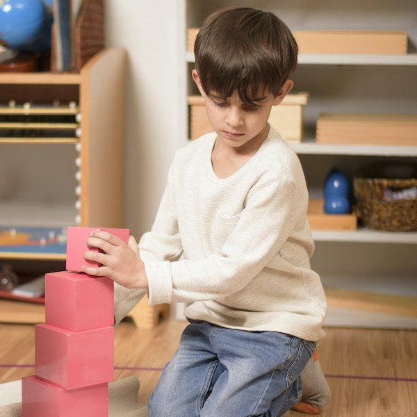 A young Montessori child builds a tower of pink blocks on a wooden floor.