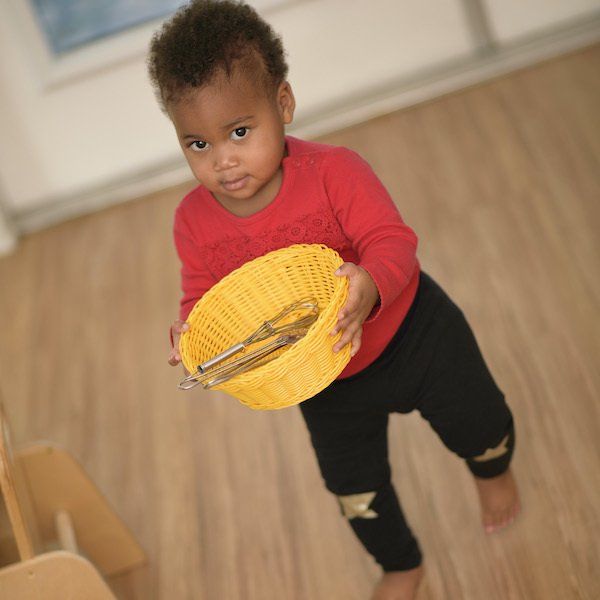 Montessori Toddler holding a yellow basket with utensils, looking at the viewer, wearing red shirt and black pants.