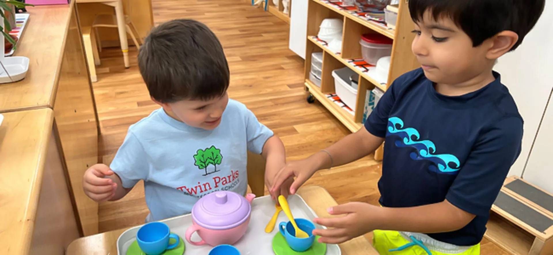 Two young children play with a toy tea set at a wooden table in a schoolroom.