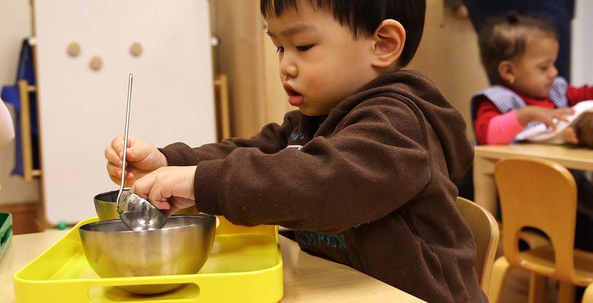 Young child in brown hoodie spoons water from bowl at a table in a classroom.
