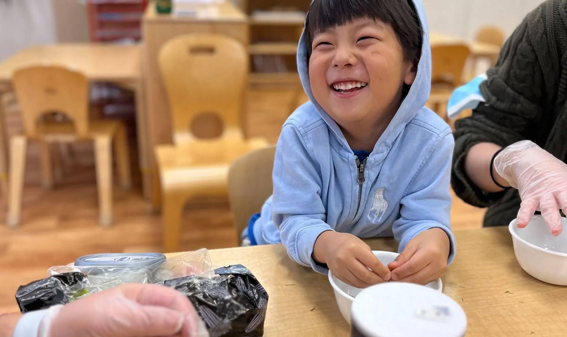 Smiling boy in blue hoodie at a table, hands near bowls, someone wearing gloves nearby.