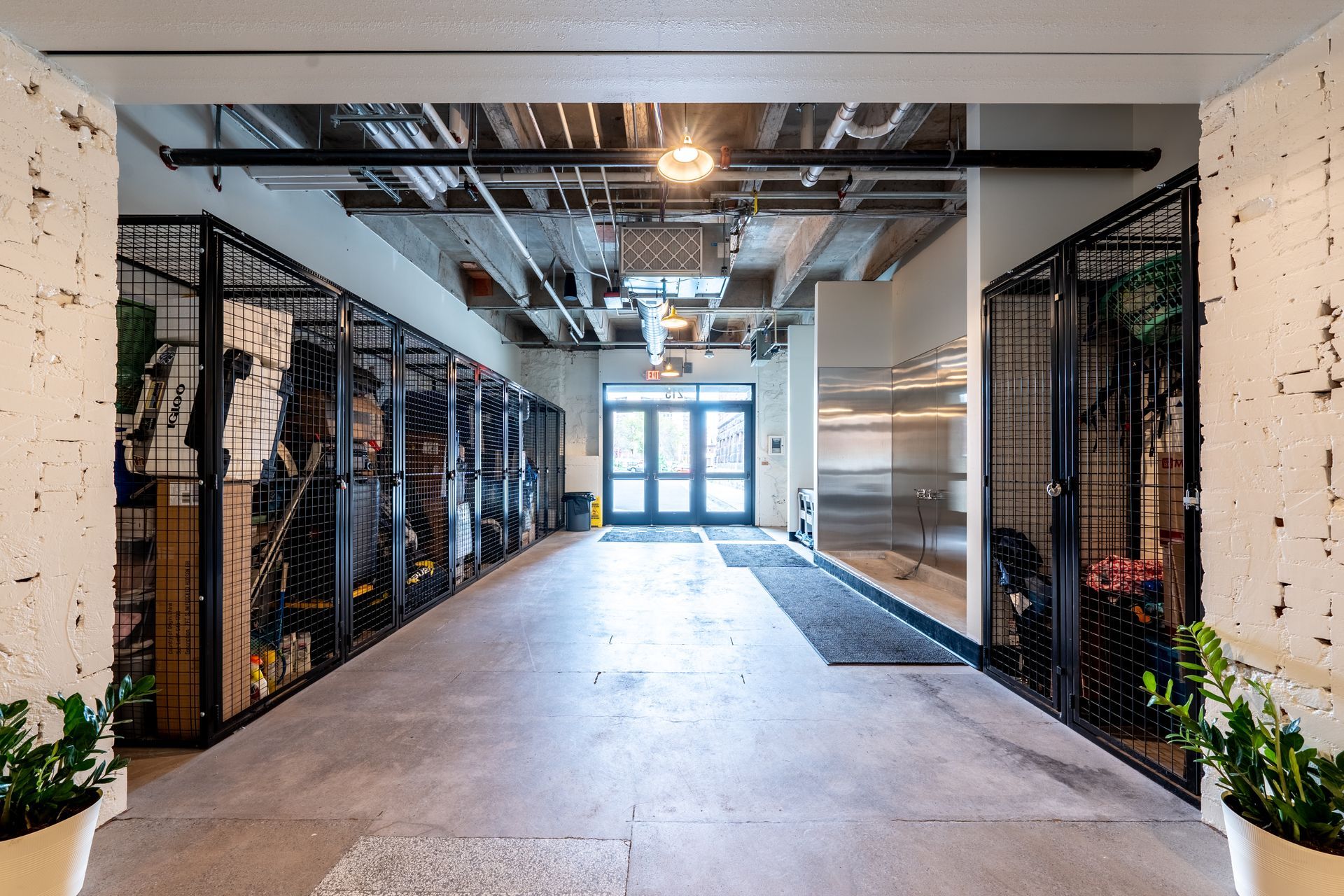 A hallway with storage lockers on both sides, leading to a doorway. Concrete floor, exposed ceiling with pipes and light fixtures.