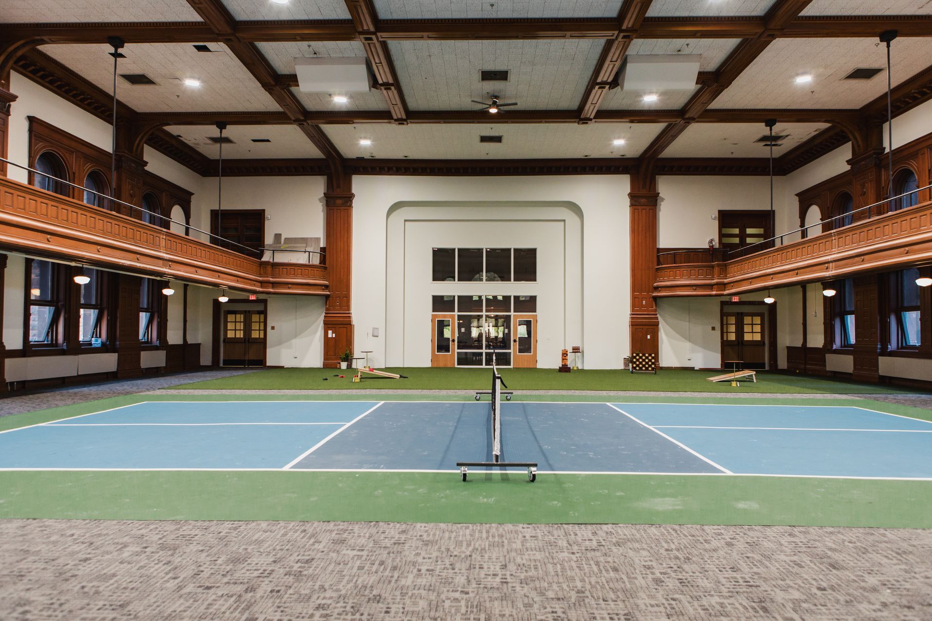 Indoor tennis court with blue and green surface, white lines, net. Red brick columns, balconies, wooden beams.