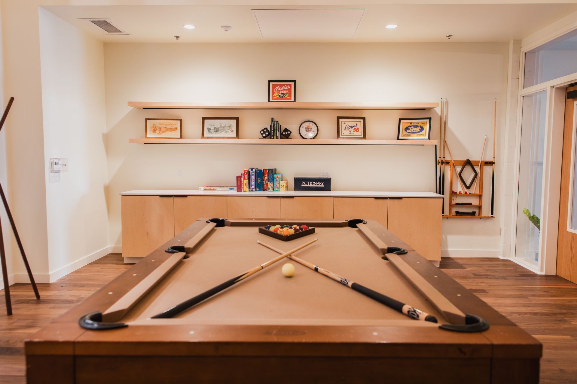 Pool table in a room with a light wood floor, shelves with decor, and a cabinet. Cue sticks crossed on the table.