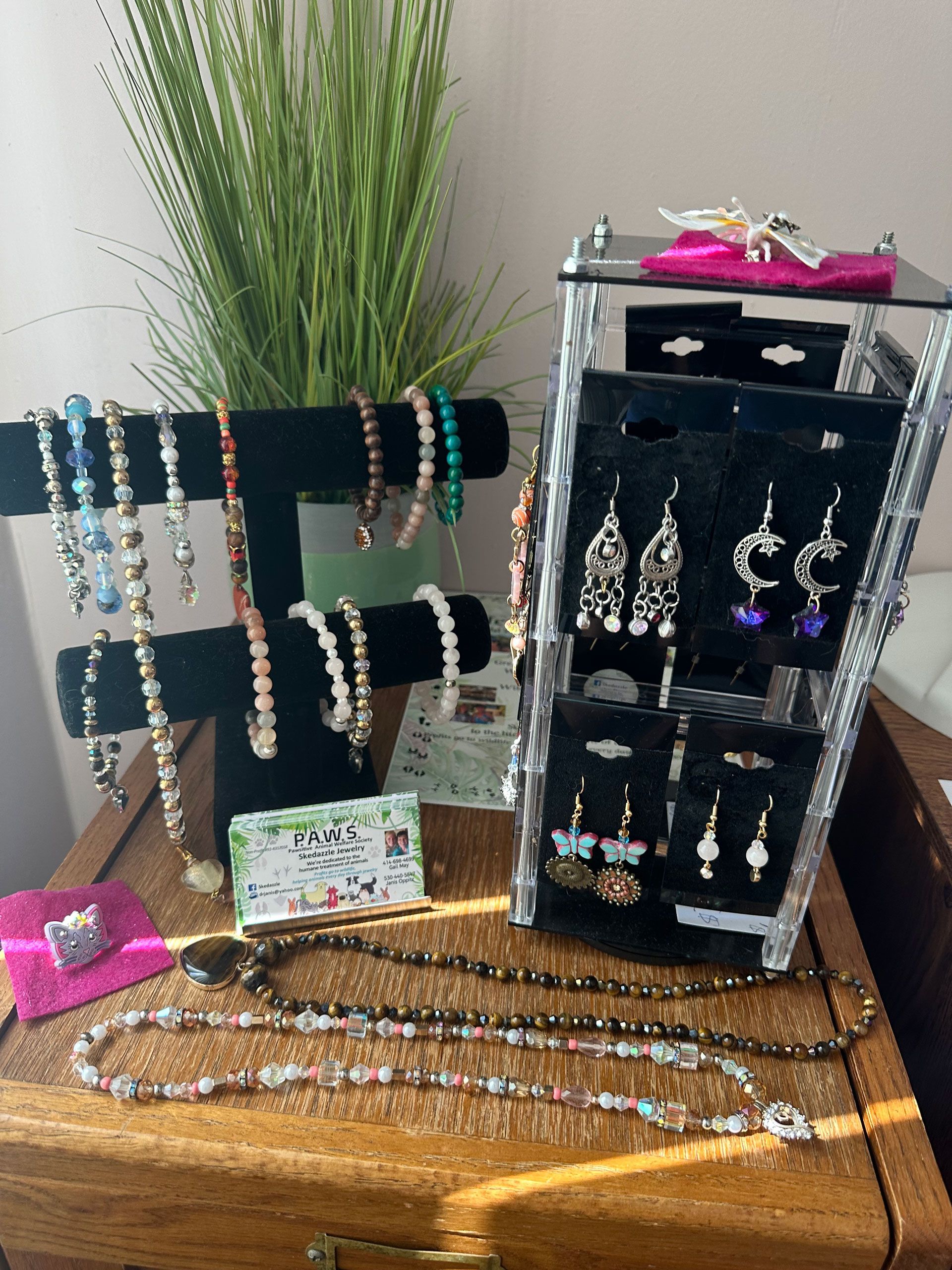 Jewelry display with necklaces, bracelets, and earrings on a wooden surface, near a plant.