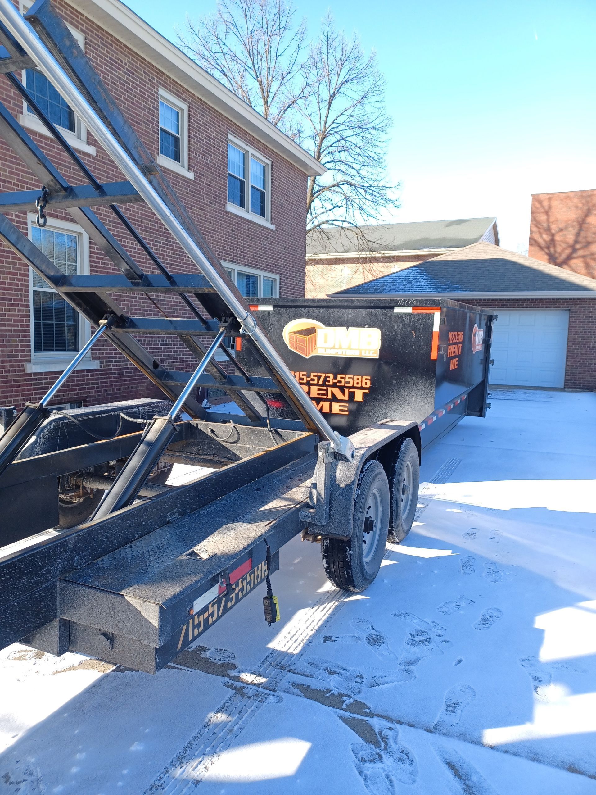 A dumpster trailer is parked in front of a brick house in the snow.