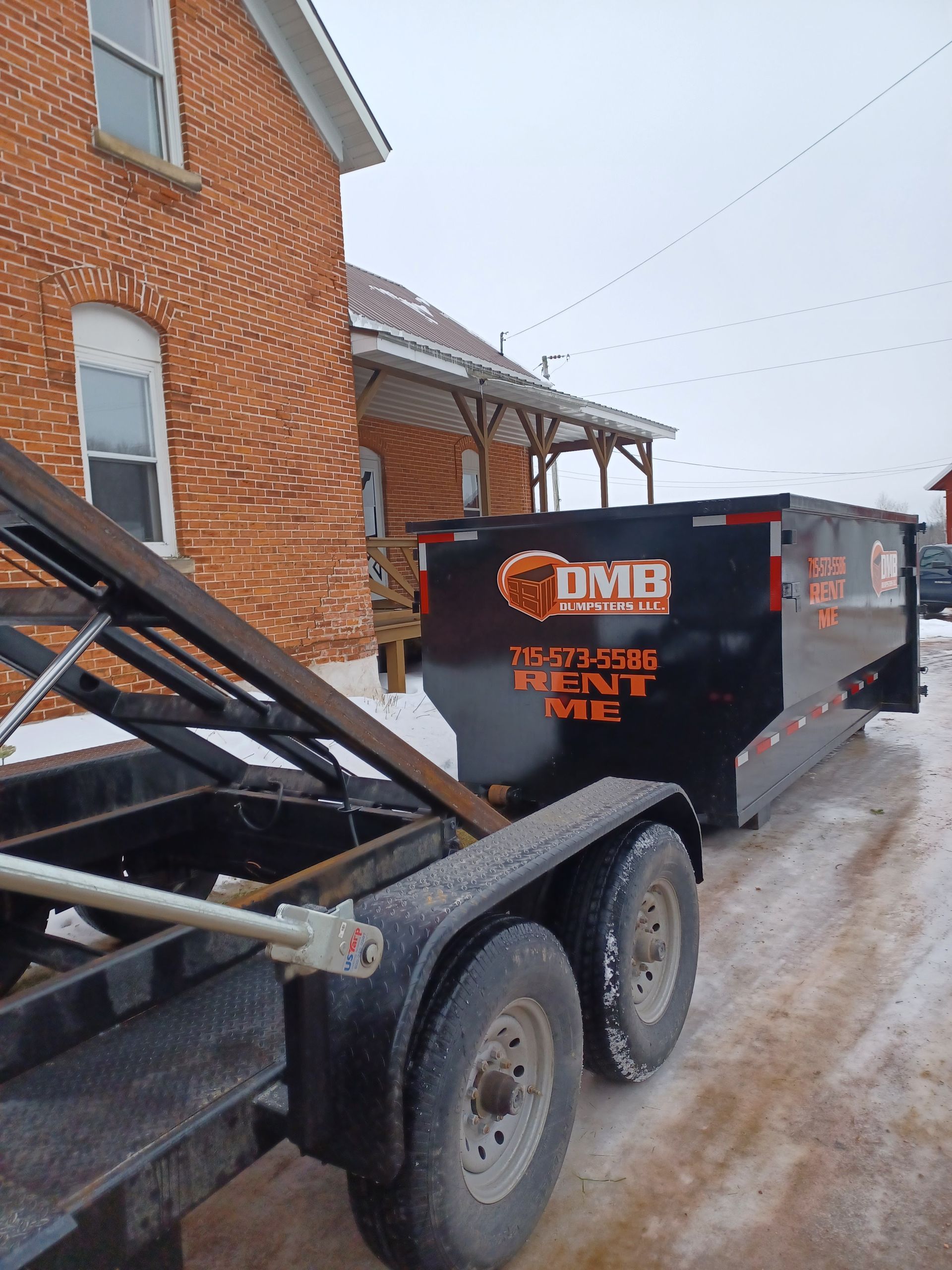 A dumpster is being pulled by a trailer in front of a brick building.