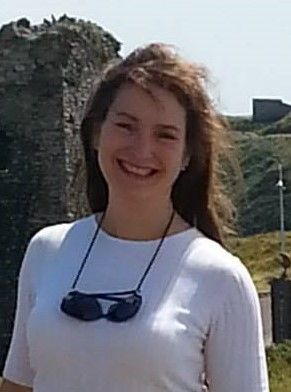 Woman with brown hair smiles in front of a stone structure, wearing a white shirt and sunglasses.