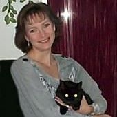 Woman smiling, holding a black cat with glowing eyes; sitting near maroon curtains and greenery.