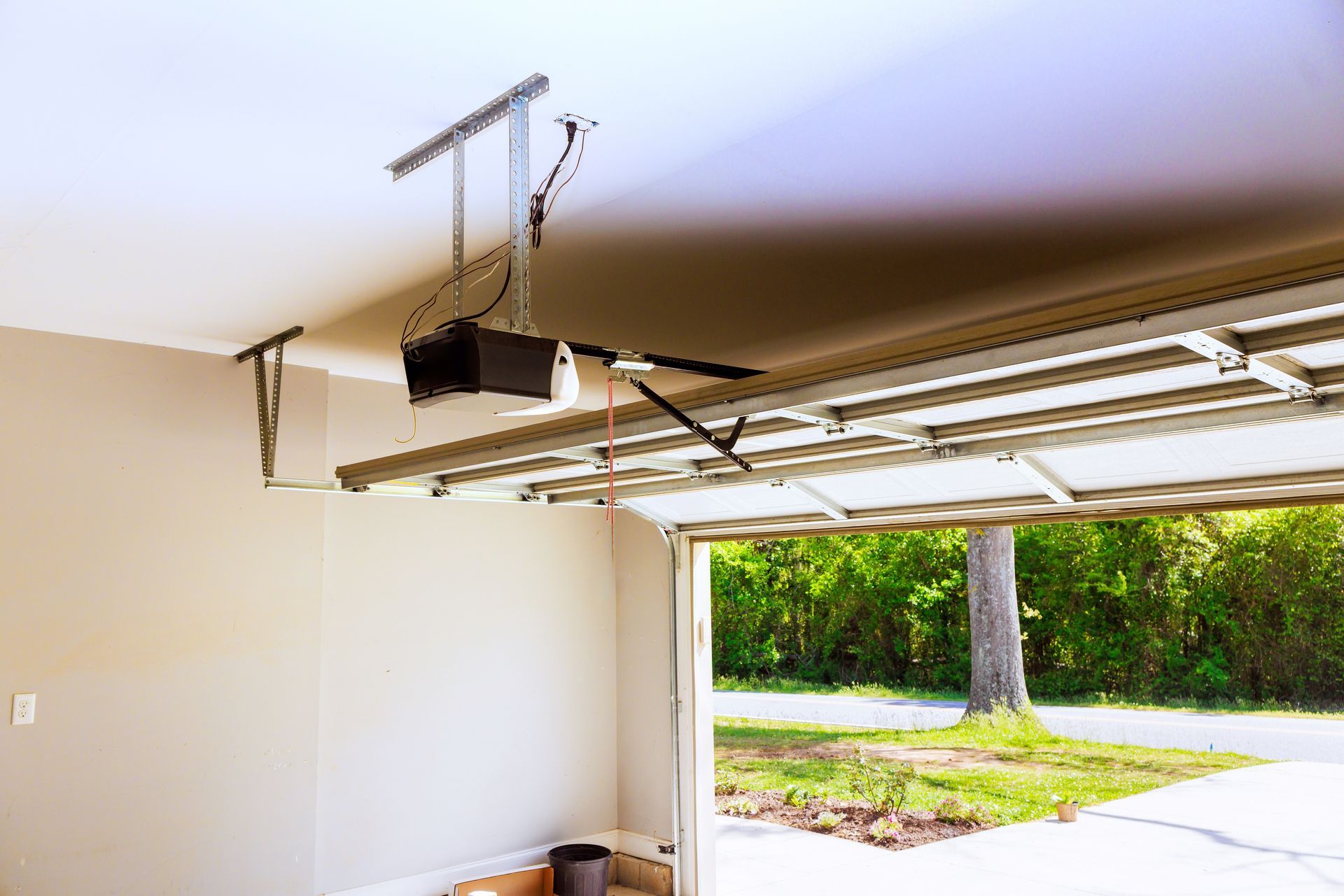 Open garage with ceiling-mounted door opener and view of driveway and green trees outside