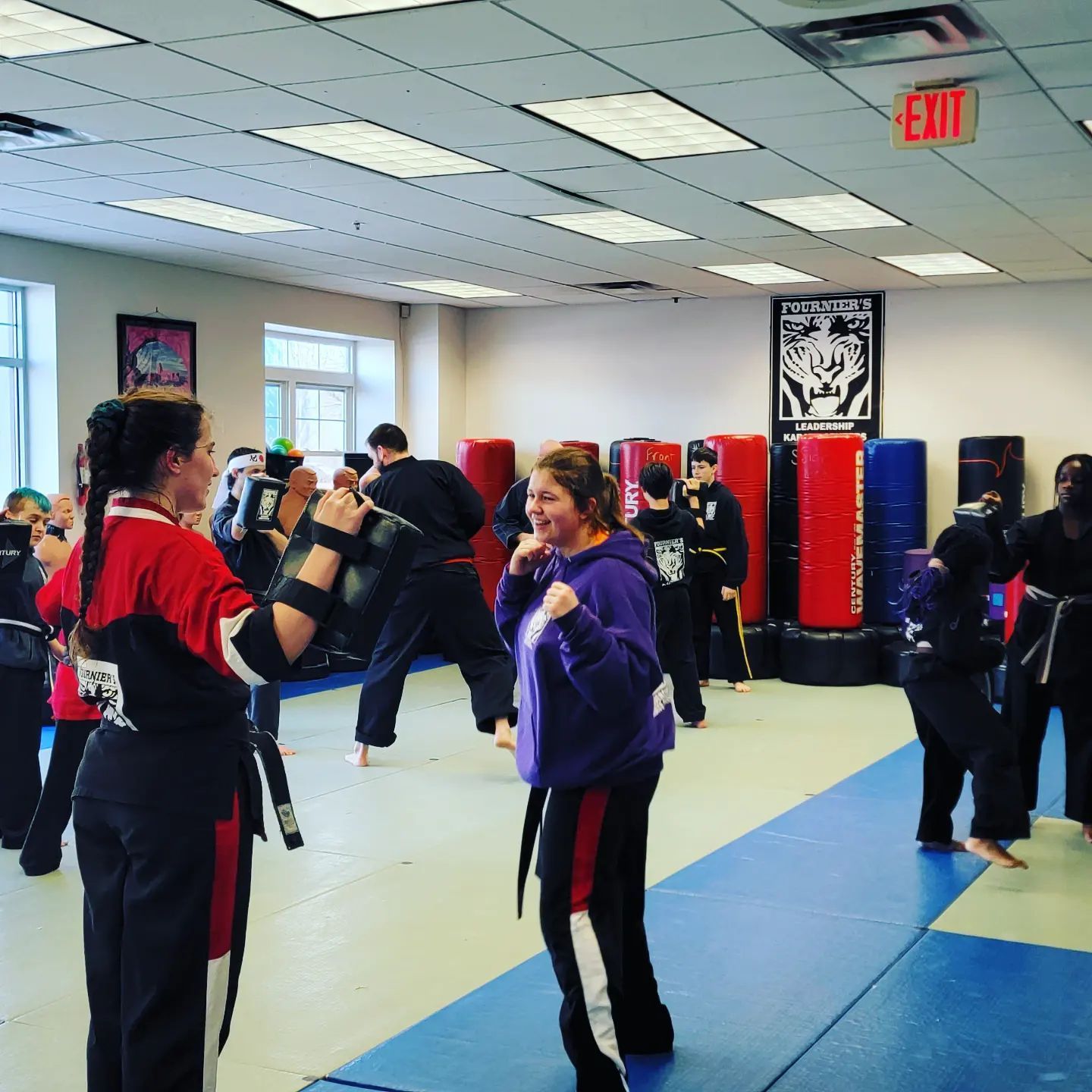 A group of people are practicing martial arts in a gym with an exit sign above them