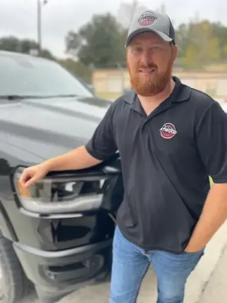 Three people in a garage discuss car detailing while applying product to a black car.