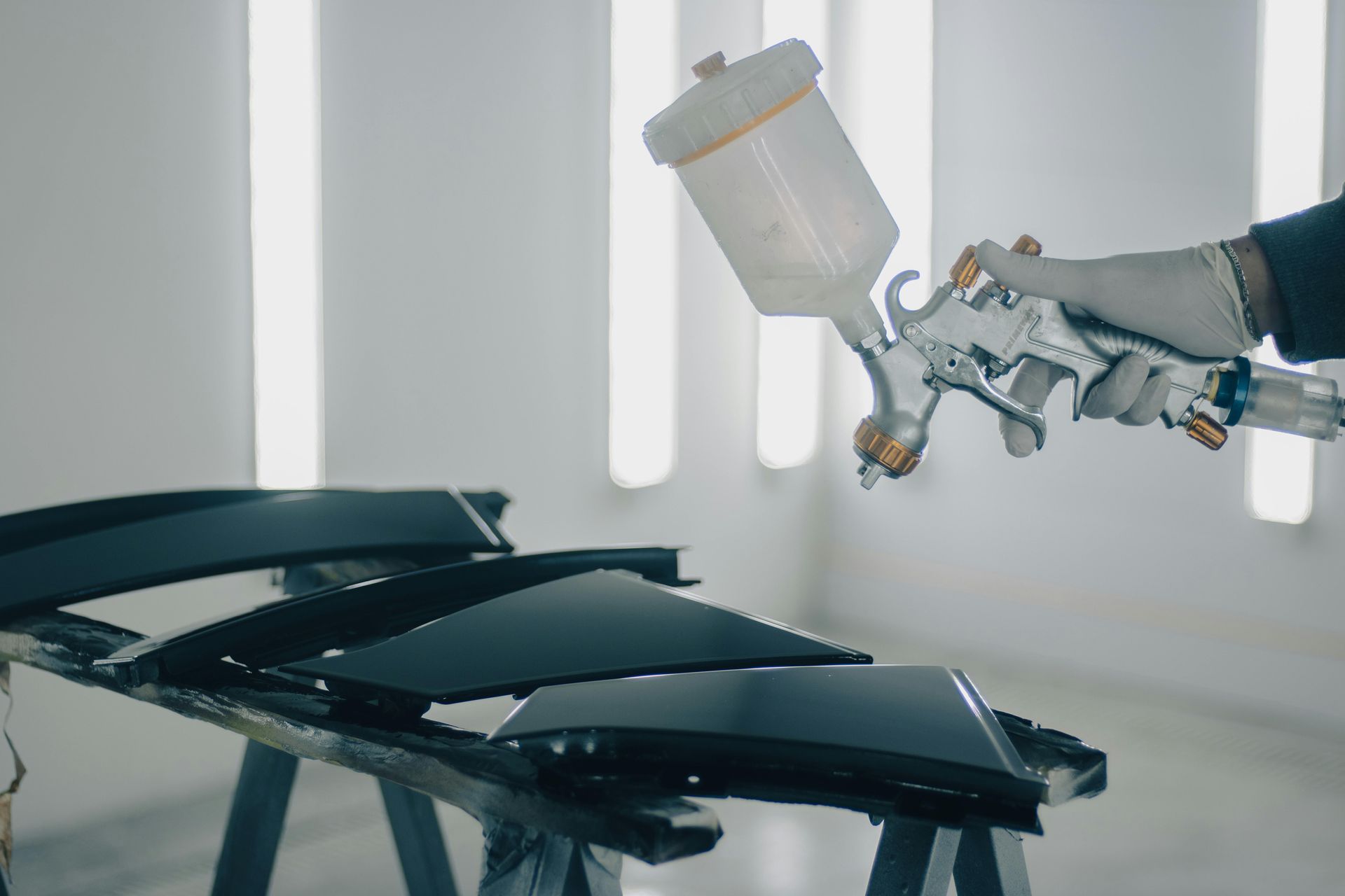 A person spray painting black car parts with a paint gun in a well-lit workshop.