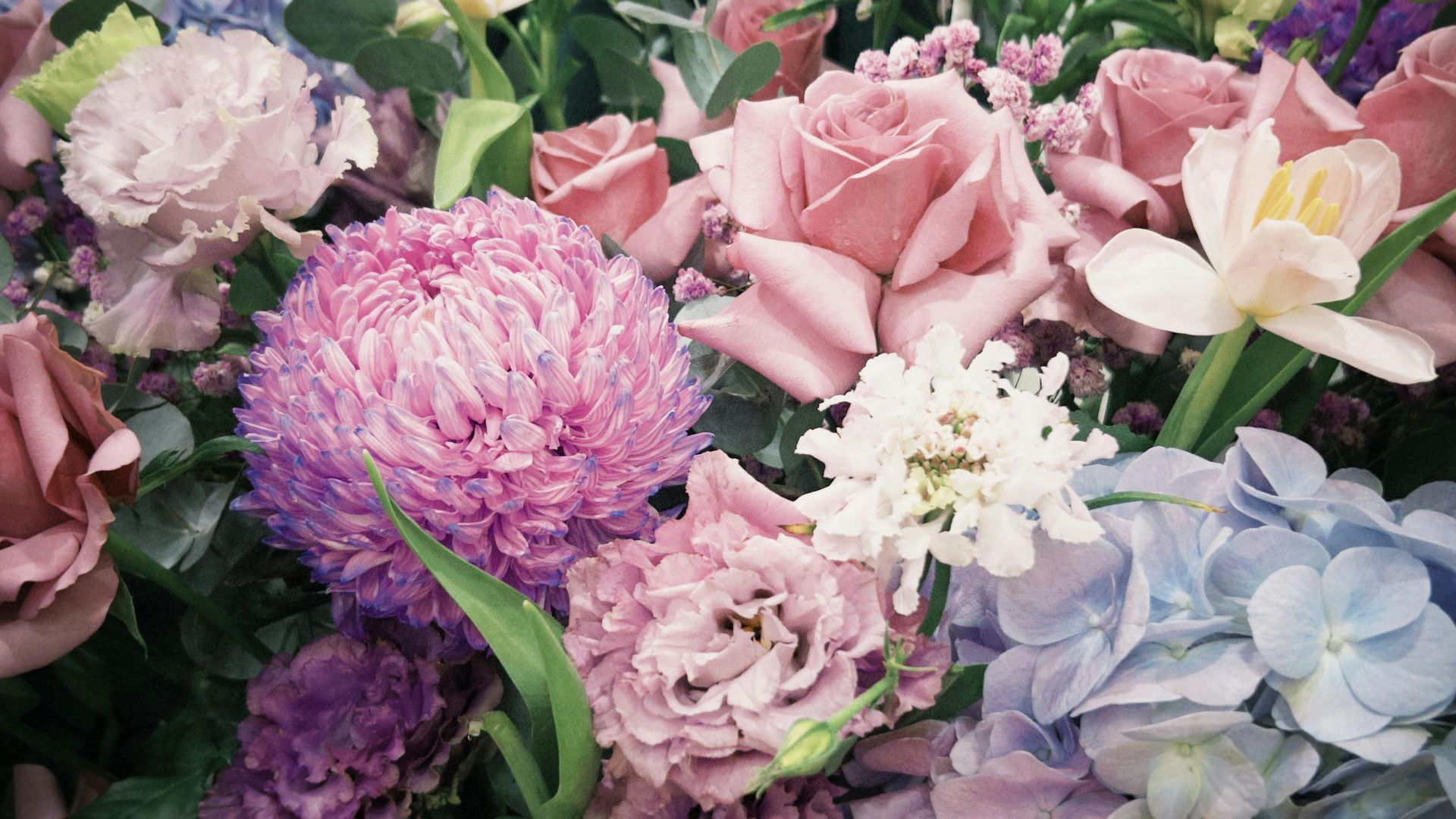 Assorted pastel flowers, including pink, purple, and blue blooms in a lush bouquet
