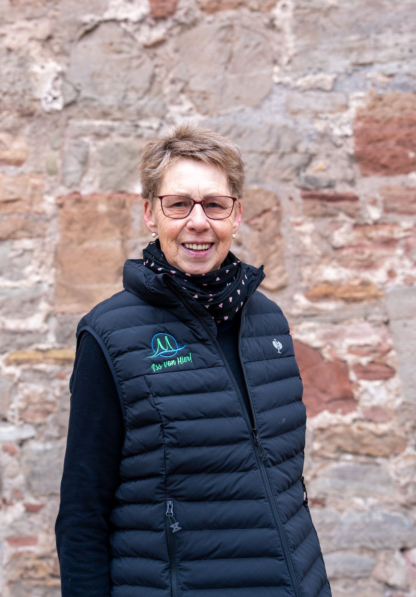Eine Frau mit Brille und schwarzer Weste lächelt vor einer Backsteinmauer.