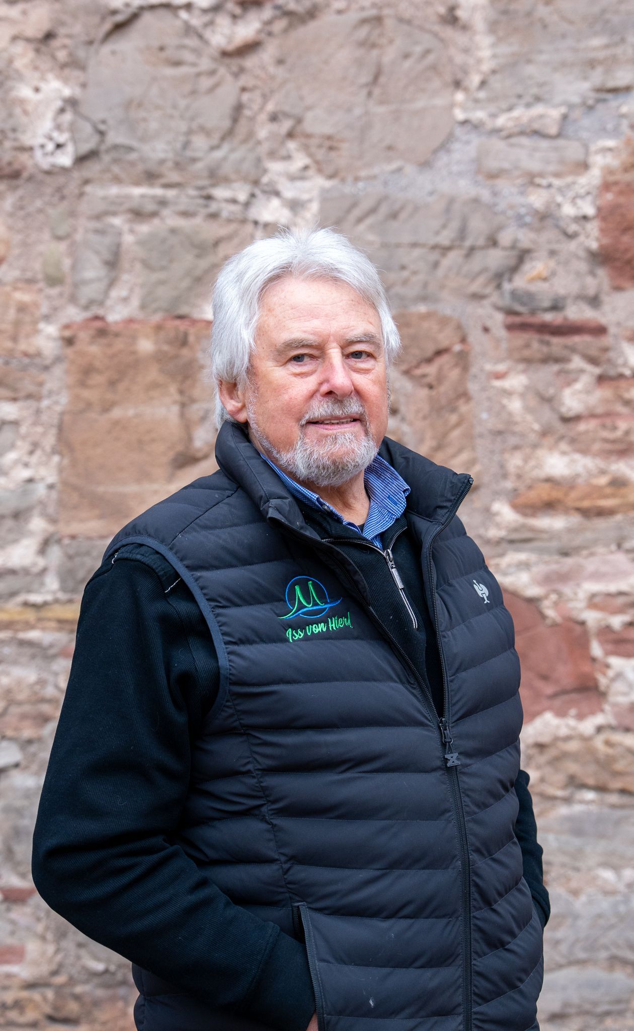 Ein Mann mit grauem Haar und Bart, der eine schwarze Weste und Jacke trägt, steht vor einer Steinmauer.