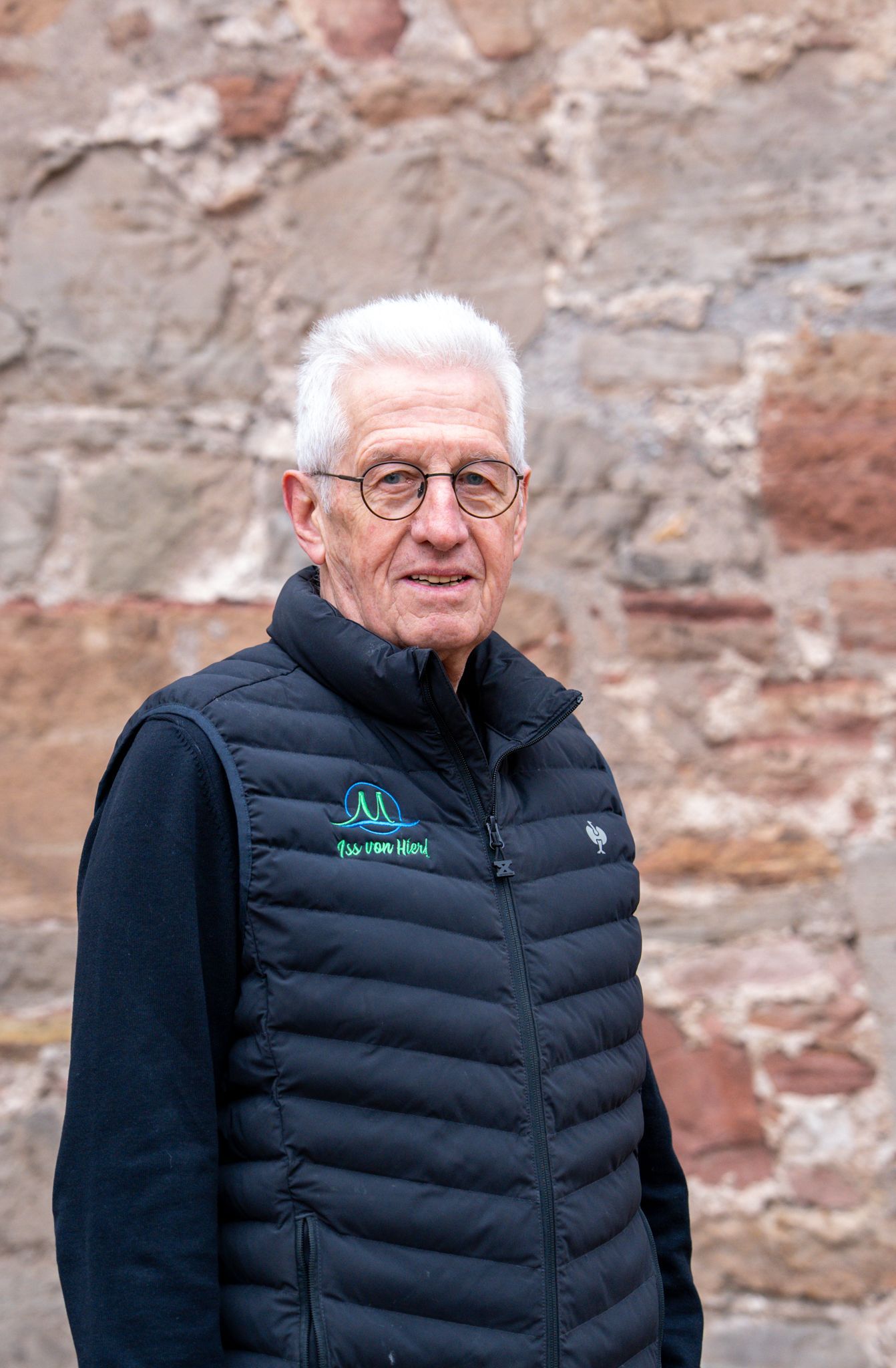Mann mit weißen Haaren, Brille und dunkelblauer Weste, steht vor einer Steinmauer.
