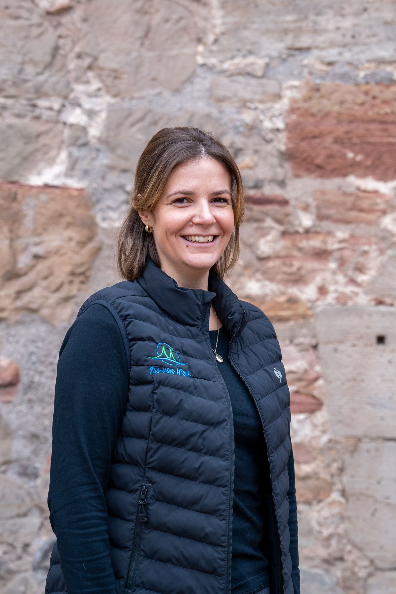 Eine Frau in schwarzer Weste lächelt vor einer Steinmauer.