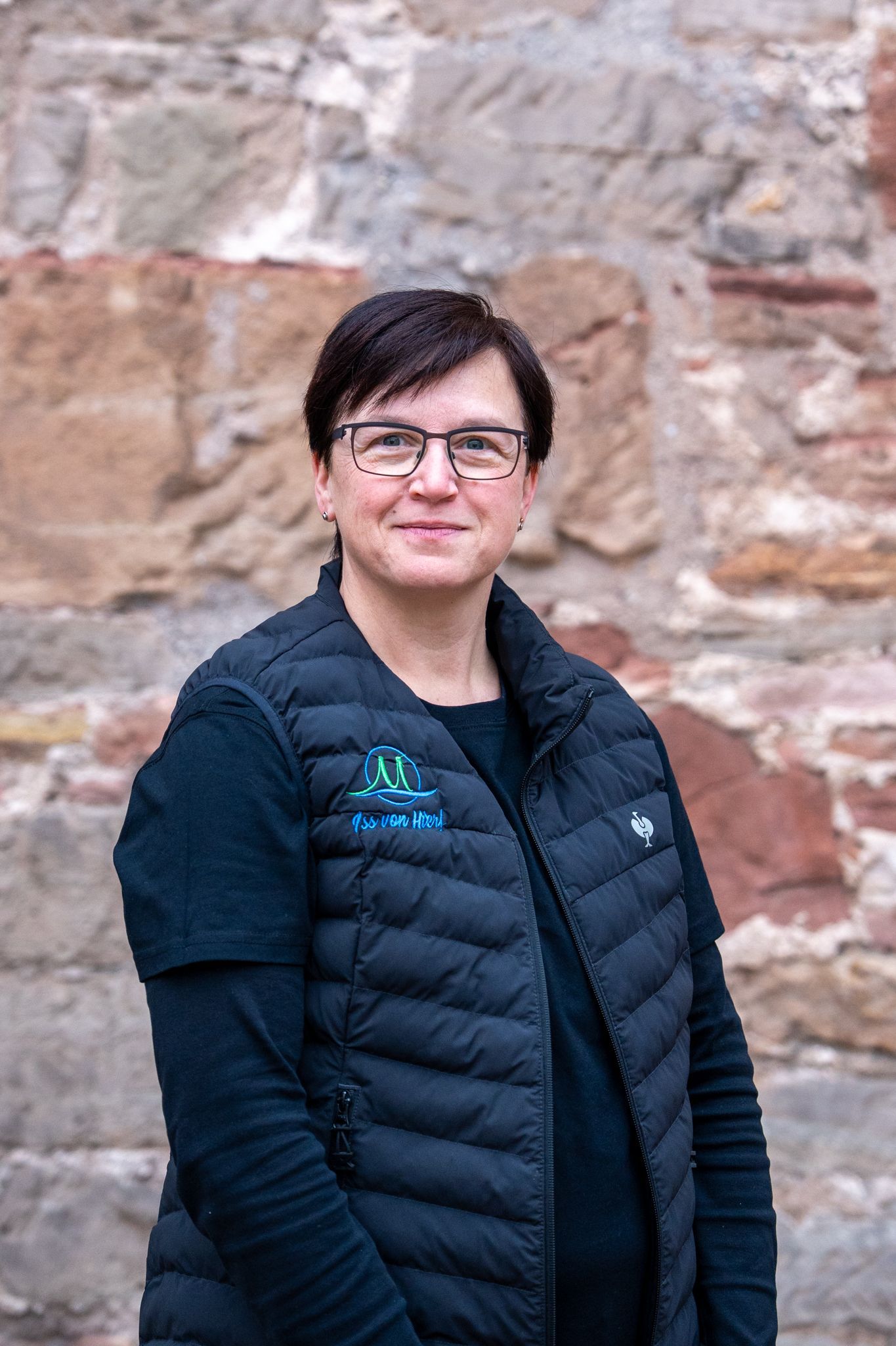 Frau mit kurzen Haaren und Brille in schwarzer Weste steht vor einer Backsteinmauer.