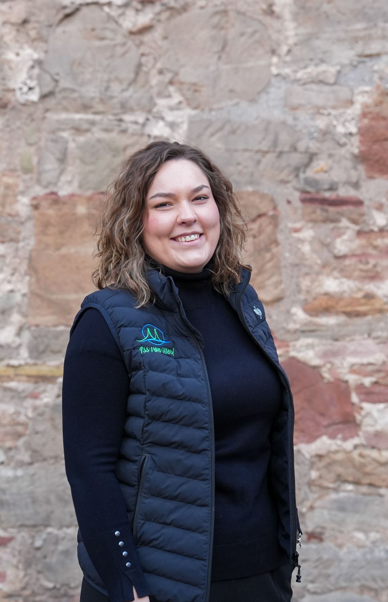 Eine lächelnde Frau in schwarzer Weste und schwarzem Rollkragenpullover steht vor einer Steinmauer.