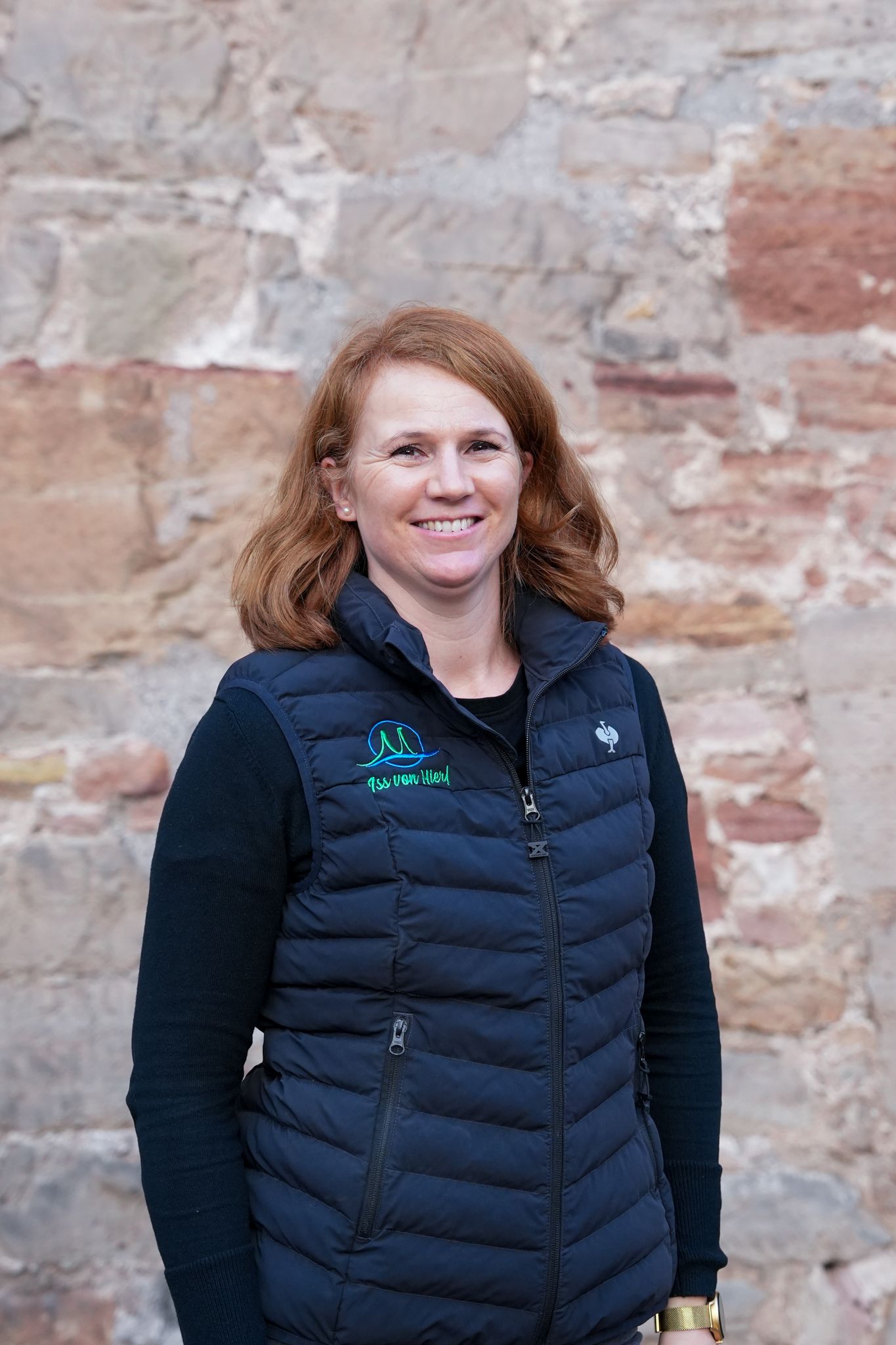Frau mit roten Haaren, lächelnd, trägt eine blaue Weste und steht vor einer Steinmauer.