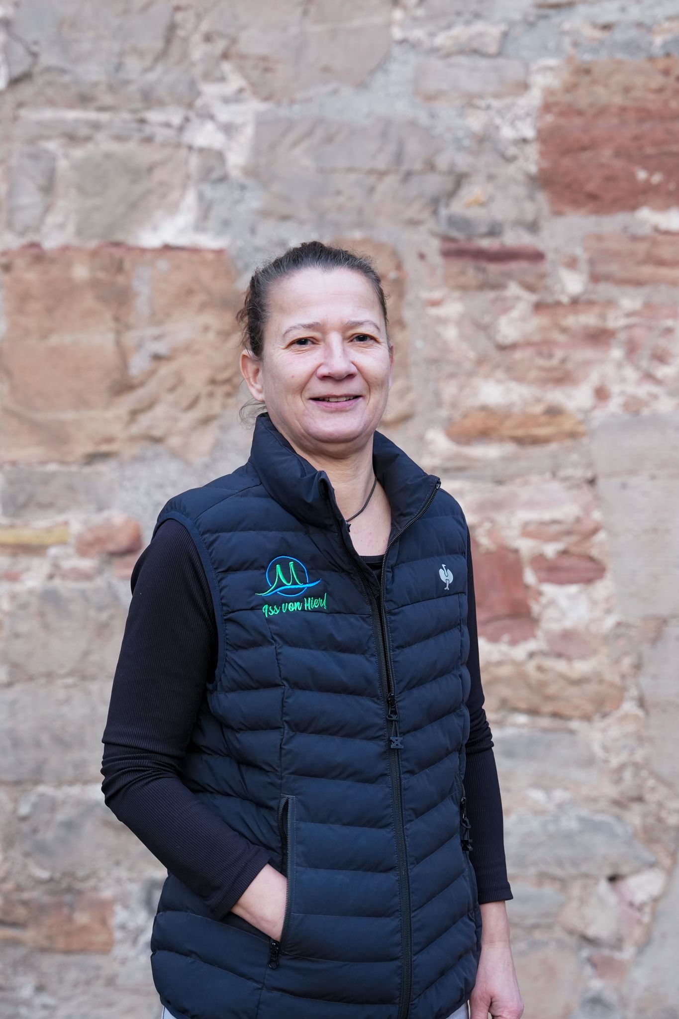 Eine Frau in einer dunkelblauen Weste lächelt und steht vor einer Steinmauer.