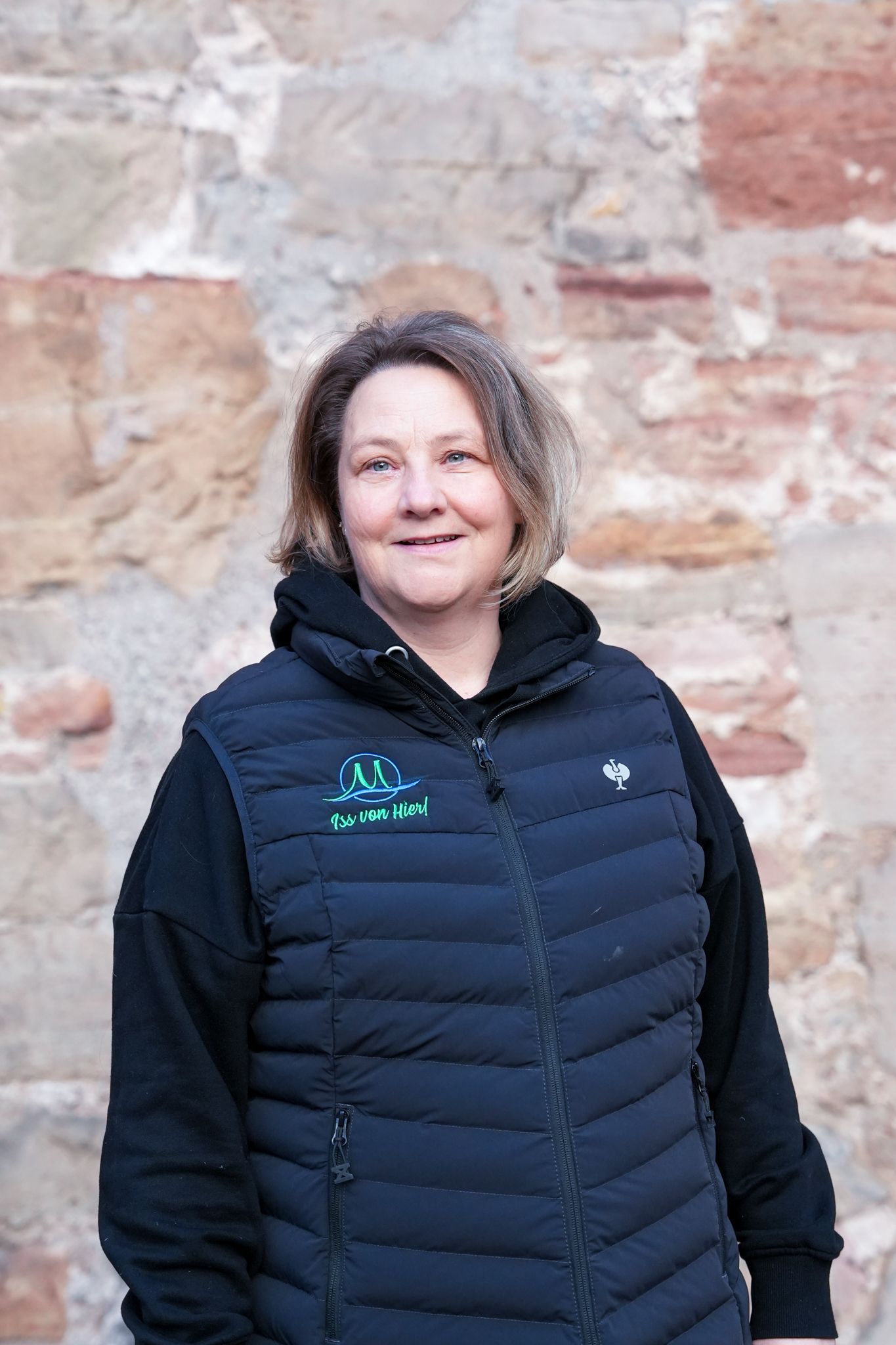 Eine Frau in schwarzer Weste und Kapuzenpulli lächelt vor einer Steinmauer.