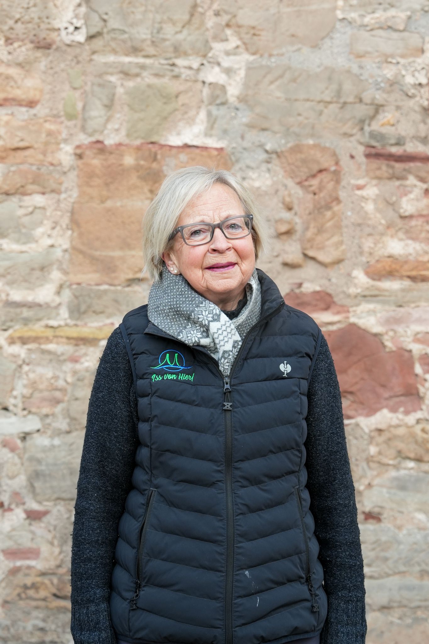 Eine Frau mit Brille und Steppweste lehnt lächelnd an einer Steinmauer.