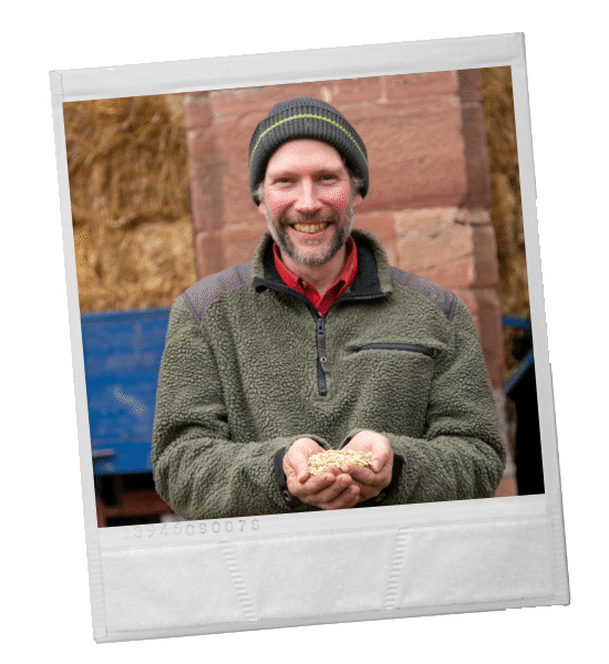 Mann mit Samen in der Hand, lächelnd, mit Hut und Fleecejacke. Im Freien, im Hintergrund Heuballen.