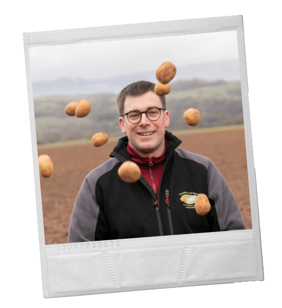 Ein Mann mit Brille lächelt, umgeben von herumfliegenden Kartoffeln auf einem Feld.