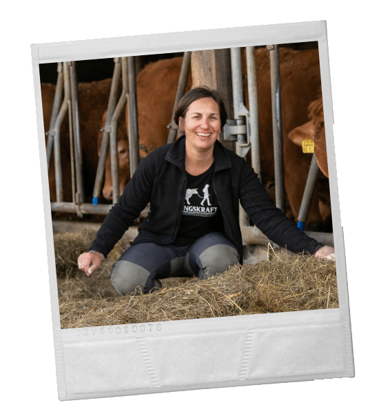 Eine Frau kniet lächelnd in einem Stall zwischen Kühen. Sie befindet sich im Heu und trägt dunkle Kleidung.