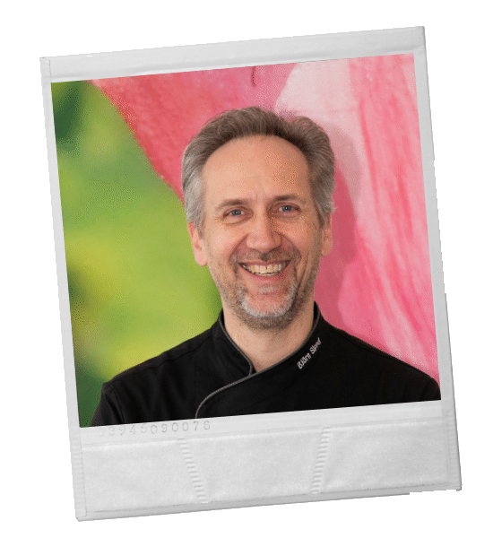 Ein lächelnder Koch in schwarzer Uniform, eingerahmt von einem weißen Polaroid, vor einem farbenfrohen rosa-grünen Hintergrund.